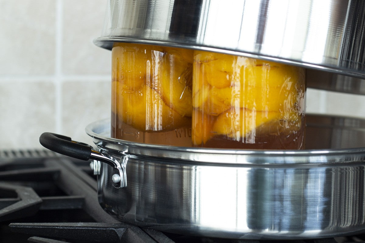 Home cook using an atmospheric steam canner to safely preserve jars of homemade jam and peaches