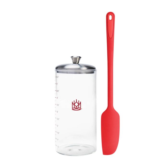 Clear container with lines to measure sourdough starter growth with silver lid and red spatula on a white background
