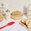 Baked goods including a loaf of bread in a bowl on a white surface with bags of flour and a red spatula.