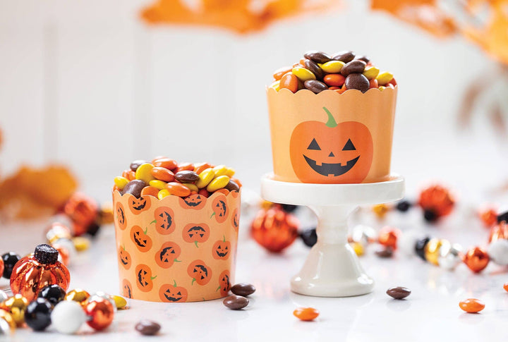 Two orange Halloween-themed candy cups filled with assorted chocolate candies, displayed on a white pedestal with scattered candy and autumn leaves in the background.