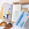 Open cookbook with a recipe for whipped butter, surrounded by wooden kitchen utensils on a marble surface.