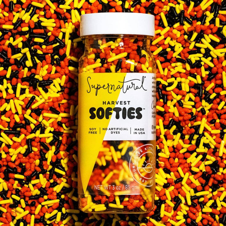 A clear plastic jar of Harvest Softies sprinkles with a white lid and a yellow-and-white label, centered on a background filled with orange, yellow, and black sprinkles.