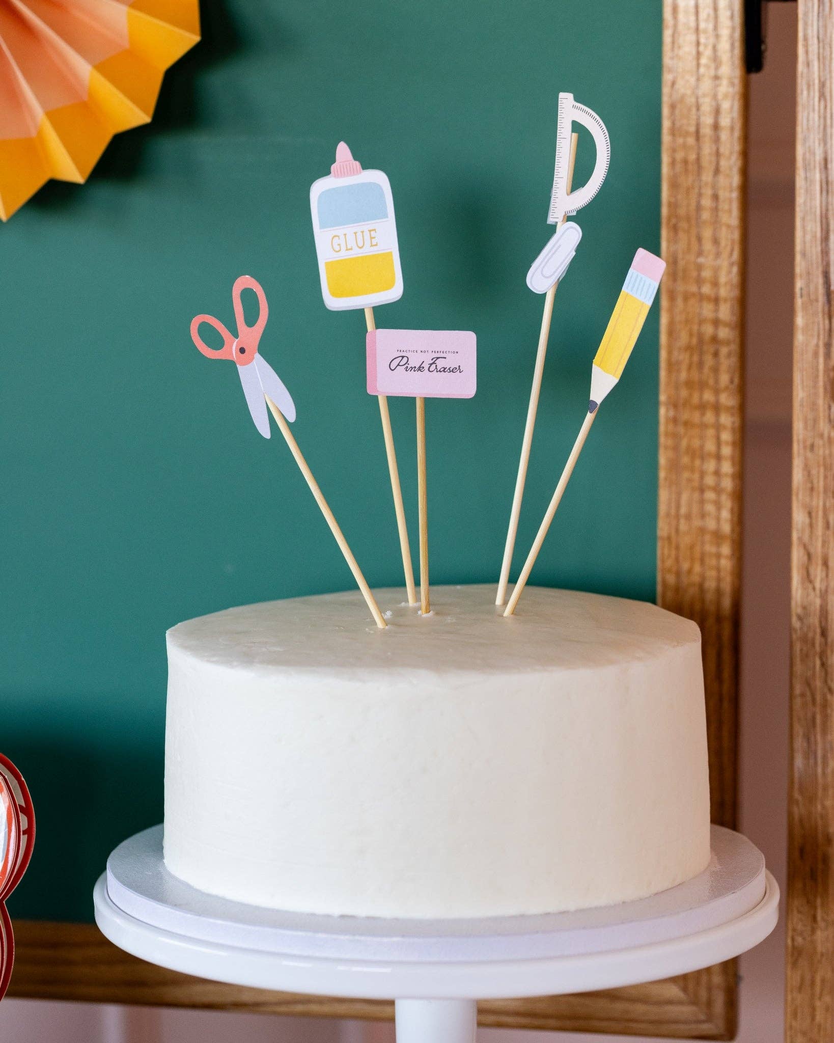 White cake with decorative toppers on a stand against a green wall.
