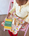 Child holding a package of pencils and a plate with sandwiches on a pink chair.
