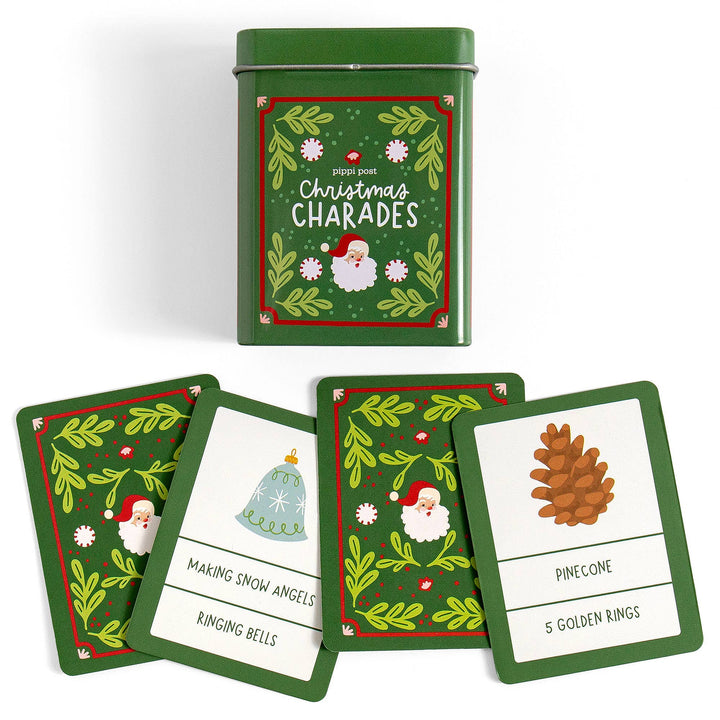 A green tin of Christmas Charades cards with several matching cards fanned out in front on a white background. The cards and tin feature a festive green border with holly leaves, red accents, Santa illustrations, and simple game words like 'Pinecone' and 'Ringing Bells'.
