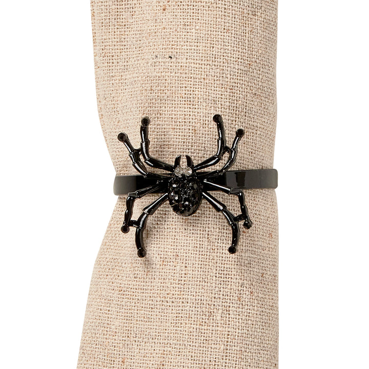 A black spider-shaped napkin ring with a glossy finish, displayed on a beige burlap-wrapped mannequin wrist against a white background.
