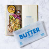 Open book showing a recipe for a dish with vegetables and a packet labeled 'The Butter Book' on a marble surface.