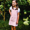 Young girl wearing a pink apron standing in a garden with greenery and flowers.