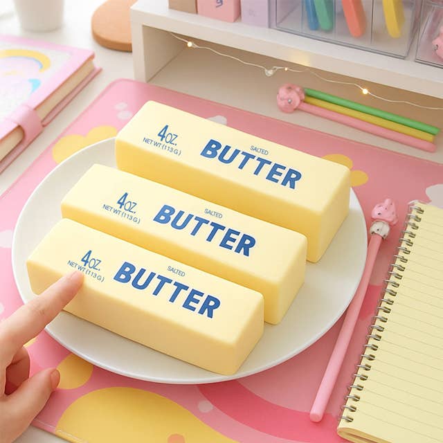 Three bars of butter squish toys on a plate with a hand pointing to one of them, set against a colorful background.