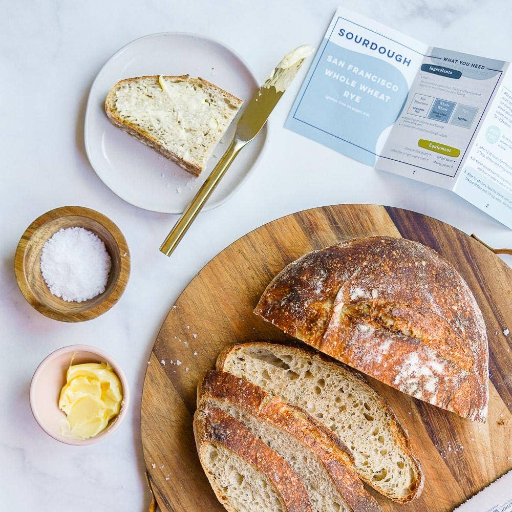 Loaves of bread on a wooden board with a slice on a plate, salt, butter, and a recipe card.