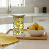 A glass canister with lemon pattern and wooden lid next to a bowl of lemons sitting on a kitchen countertop.
