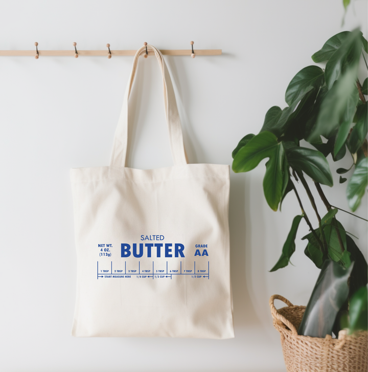 Beige tote bag with blue text on a white wall with a plant in the background
