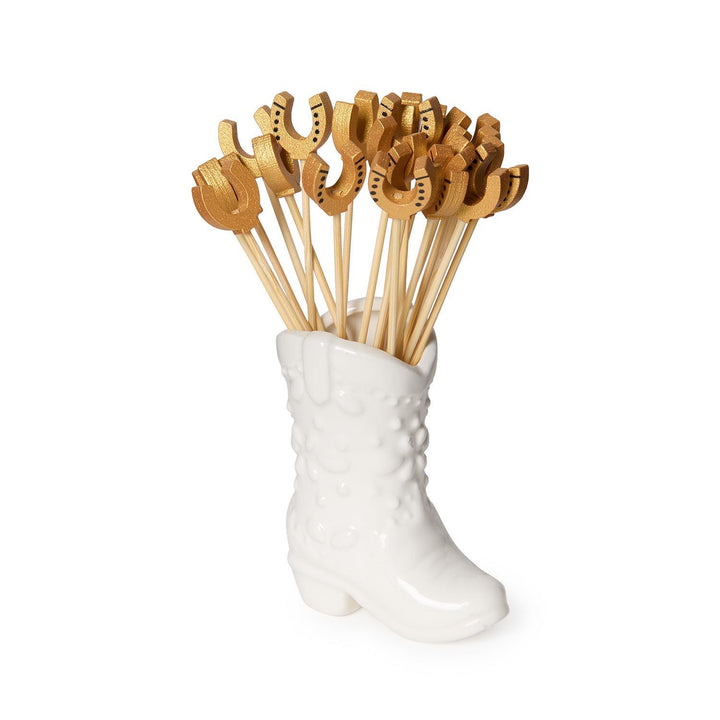 White boot-shaped holder with gold horseshoe-shaped cocktail picks on a white background