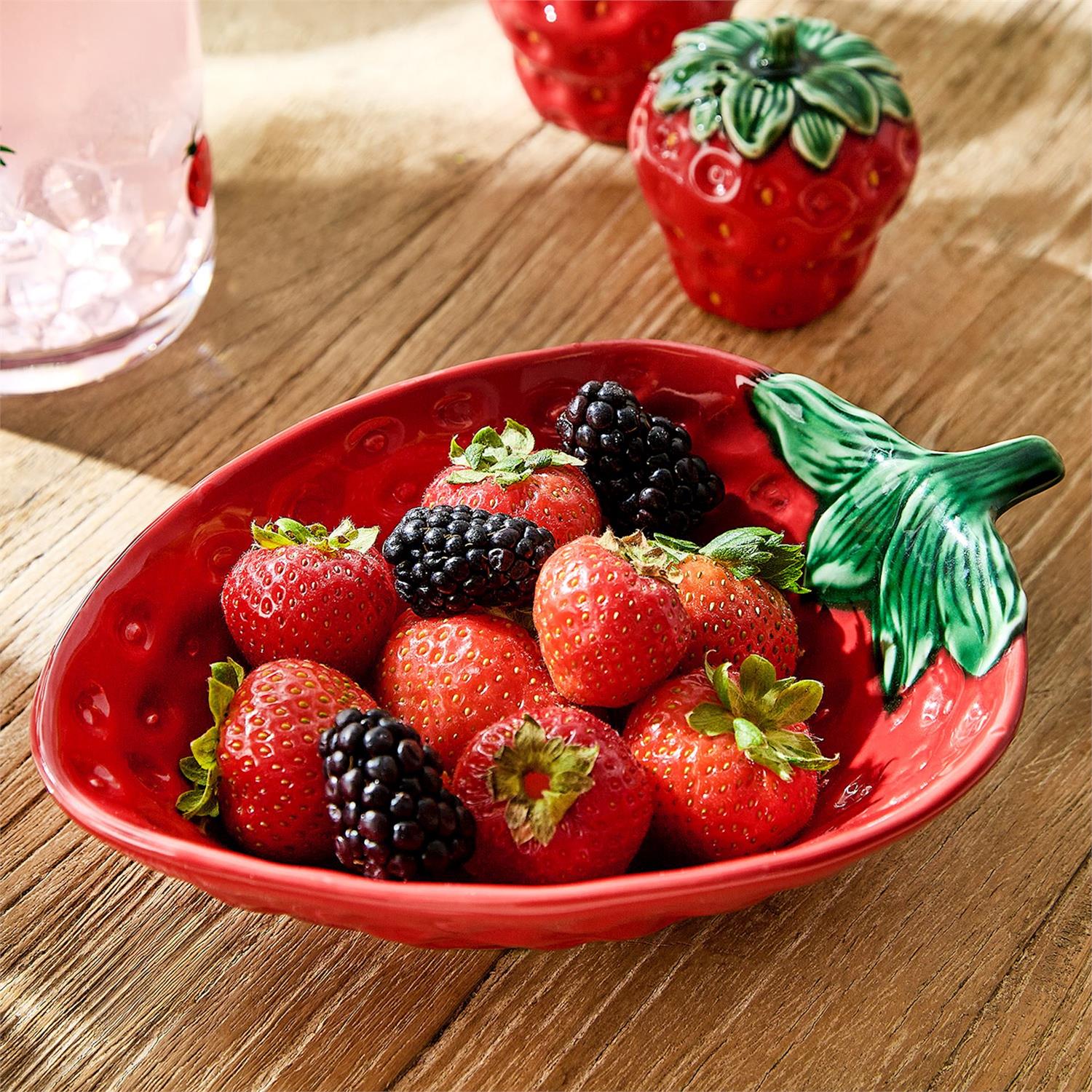 Red strawberry-shaped bowl filled with strawberries and blackberries on a wooden surface.
