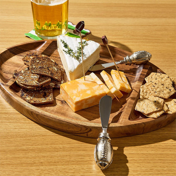 Wooden football-shaped cheese board with assorted cheeses, crackers, and a glass of beer on a wooden table.