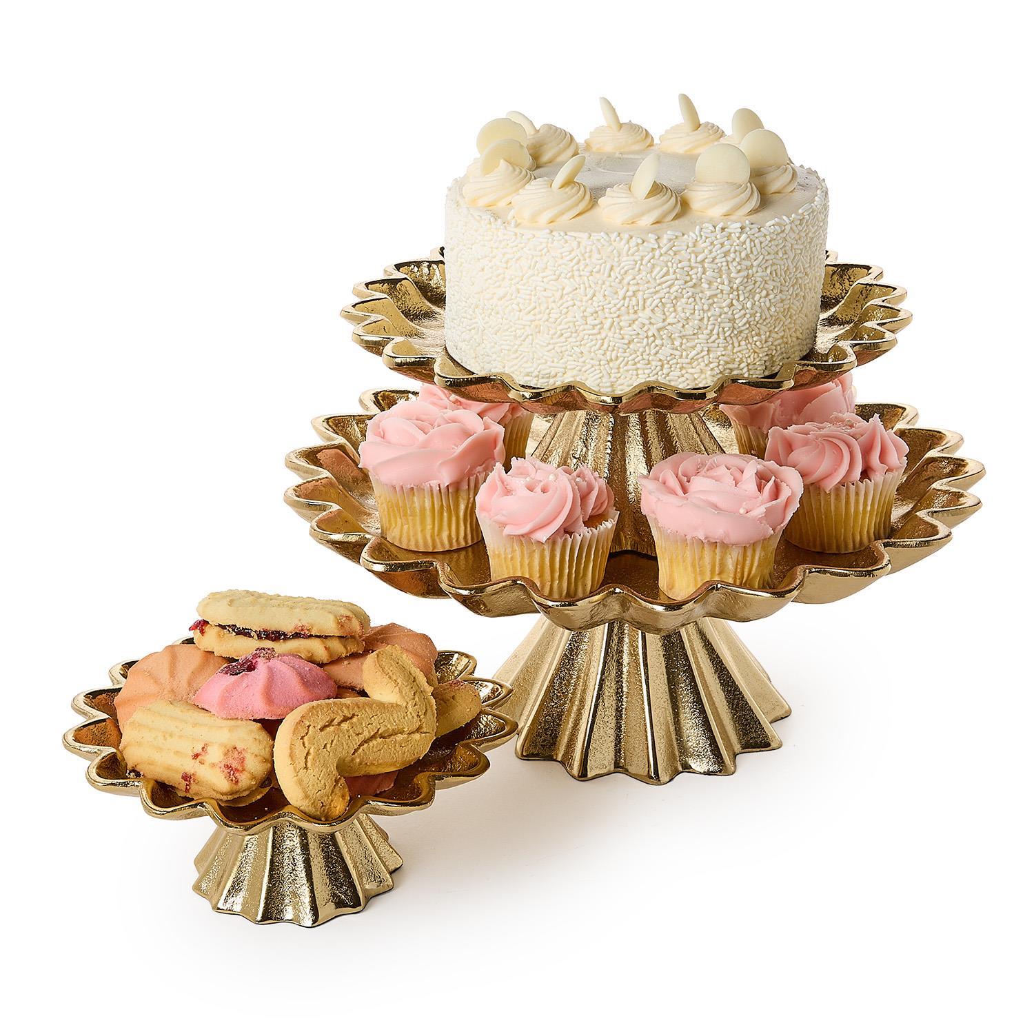 Gold cake stands with scalloped edges in three sizes with a cake, cupcakes and cookies on a white background.