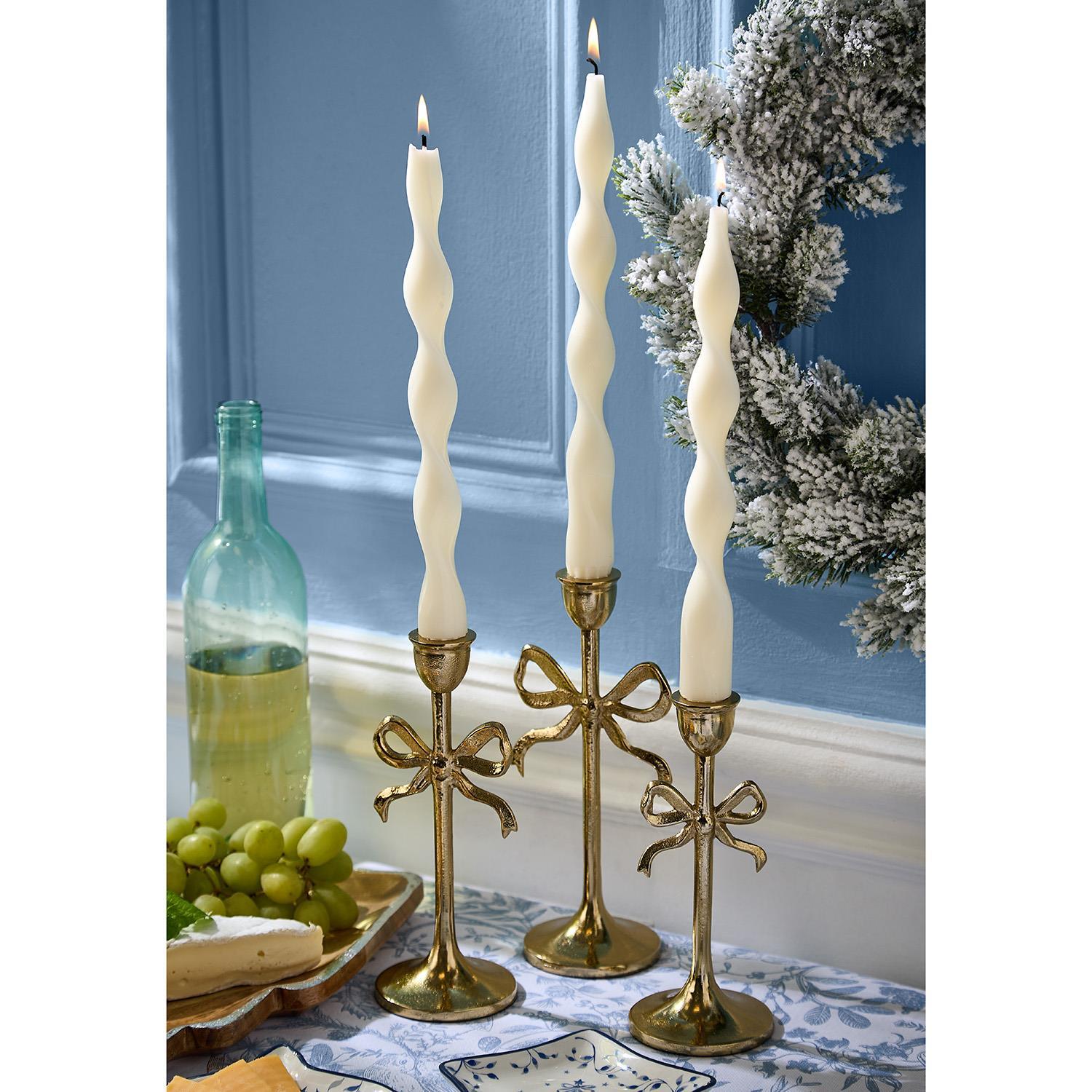Three gold candle holders with bow design holding white candles on a table with a blue wall and decorative elements in the background.