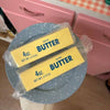 Two packages of butter squish toys on a blue plate with a pink background