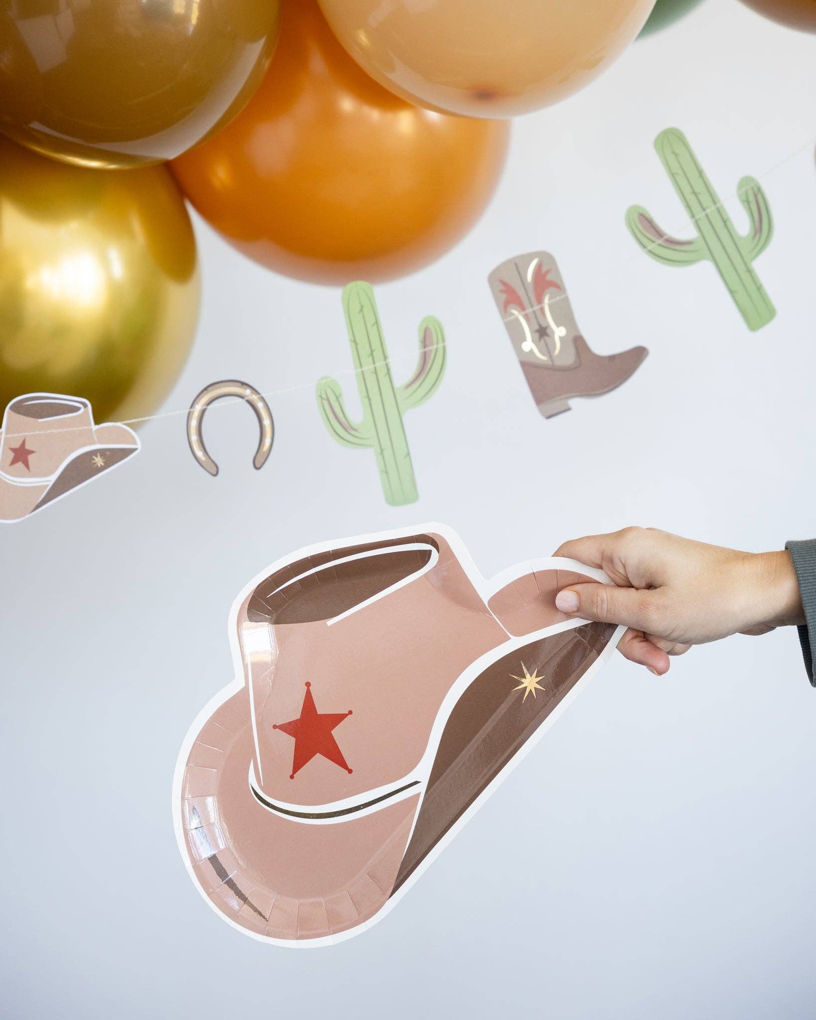 Hand holding a pink cowboy hat plate with a star, surrounded by balloons, cactus and boot decorations on a light gray background.