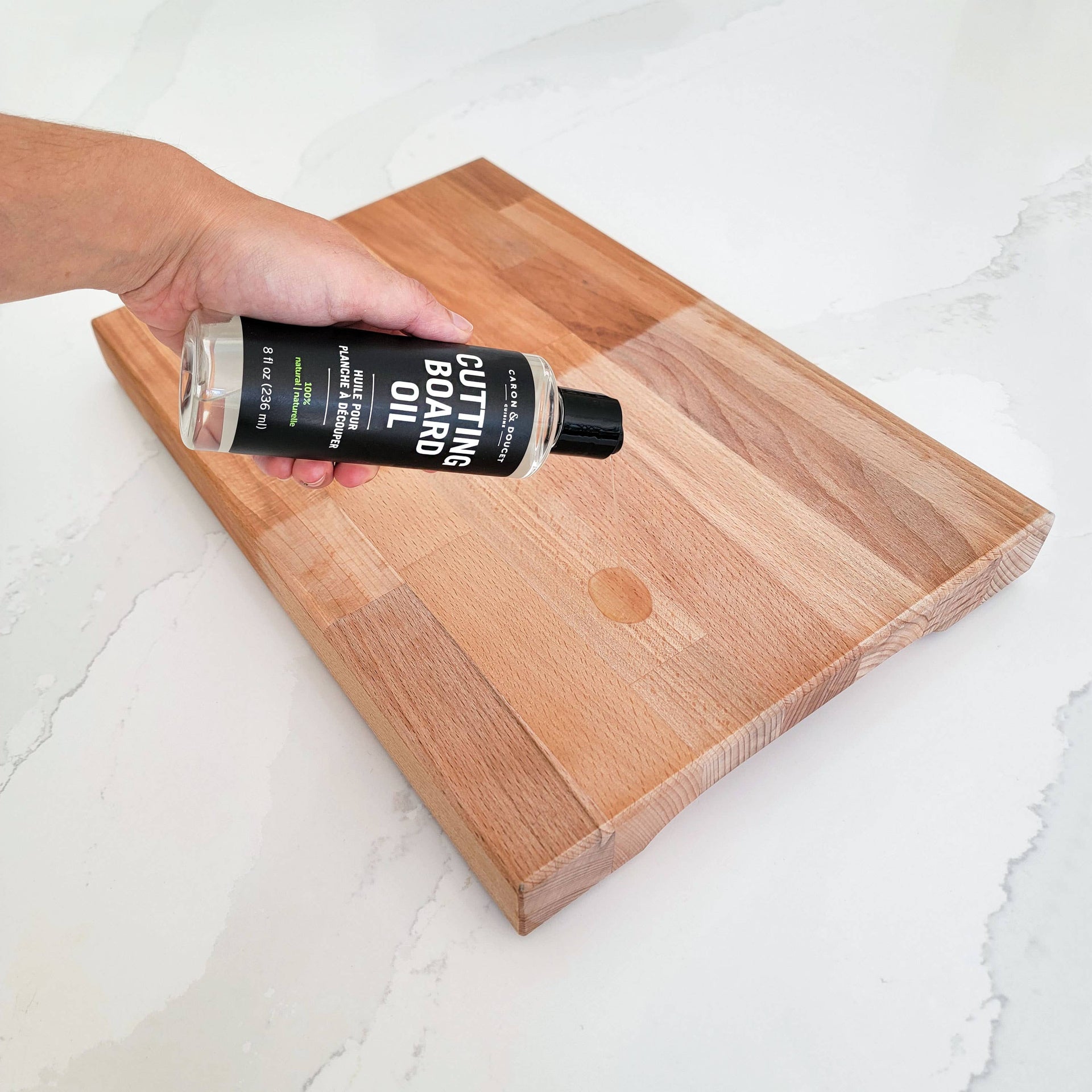 Hand holding a bottle of cutting board oil over a wooden cutting board on a white surface