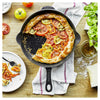 Tomato galette in a Staub skillet on a wooden table with ingredients and a plate of food.
