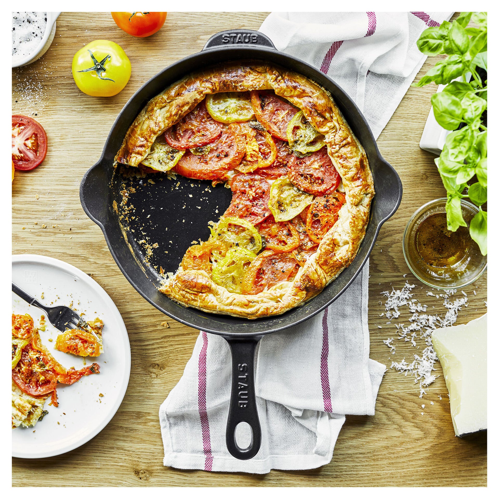 Tomato galette in a Staub skillet on a wooden table with ingredients and a plate of food.