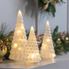 Three lit Christmas Trees on a decorative surface with greenery in the background