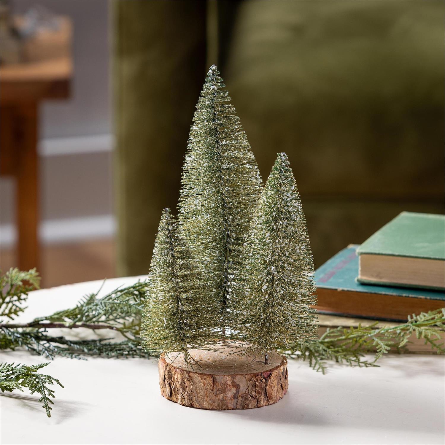 Three decorative Christmas trees on a wooden base with snow and greenery.