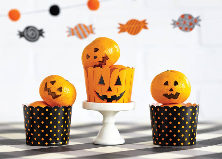 Three orange cupcakes decorated with carved jack-o'-lantern oranges, styled on a white pedestal and black-and-orange polka dot wrappers against a white background with Halloween decorations.