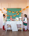 Two children in a classroom setting with a green chalkboard and decorative fans.
