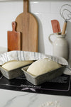 Two loaves of bread in loaf pans on a black dough mat with baking tools in the background.