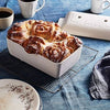 A white rectangular ceramic bread pan filled with a baked cinnamon roll or bread loaf, styled on a cooling rack with coffee mugs, a plate, and a cloth on a blue tablecloth.