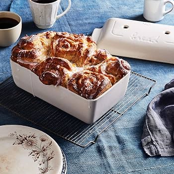 A white rectangular ceramic bread pan filled with a baked cinnamon roll or bread loaf, styled on a cooling rack with coffee mugs, a plate, and a cloth on a blue tablecloth.