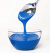 Spoon pouring blue frosting into a clear glass bowl on a white background