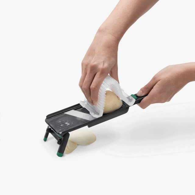 Person using a mandoline to slice a potato on a white background