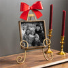 Decorative setup with a gold easel holding a black and white photo, red bow, and two red candles in gold candlesticks on a wooden surface.