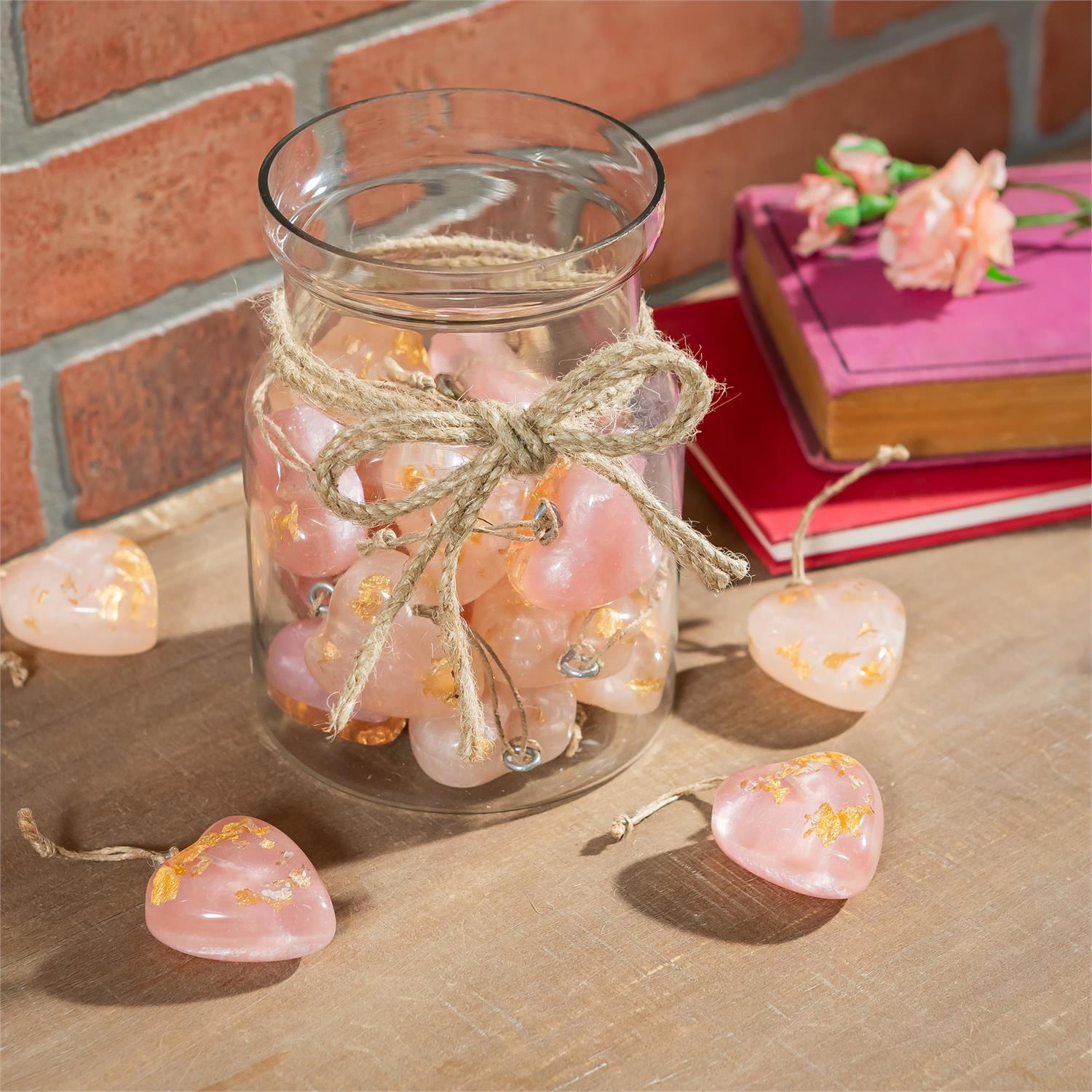 A glass jar of decorative pink hanging resin hearts sitting on a wood surface next to a stack of books in front of a brick wall.