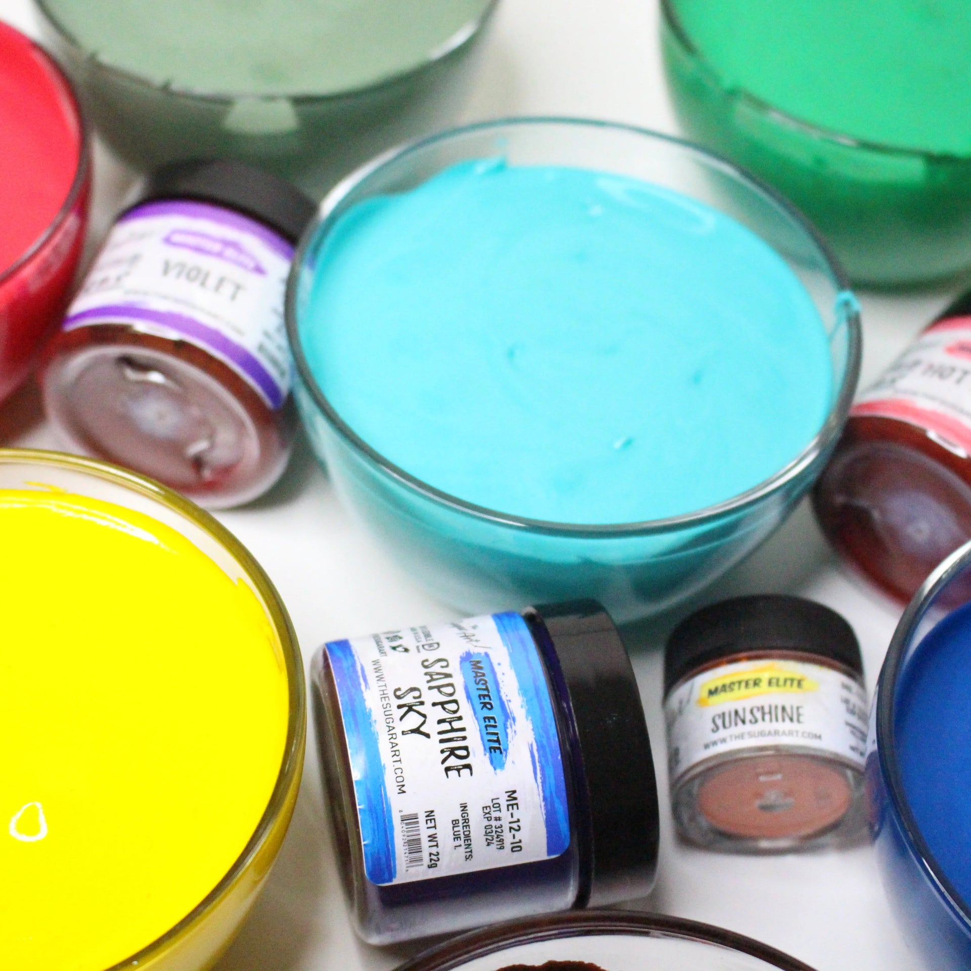 Assorted jars of powdered food color with bowls of colorful frosting on a white background 