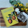 Yellow recipe book with peach design on a wooden cutting board with flowers and chocolate candy.