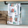 Decorative kitchen towel with winter village design hanging on a rack against a tiled wall.