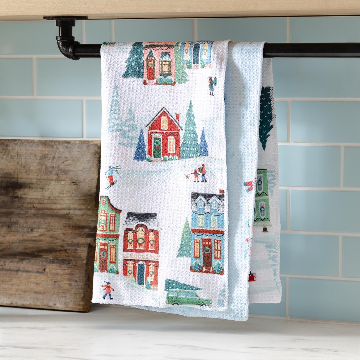 Decorative kitchen towel with winter village design hanging on a rack against a tiled wall.