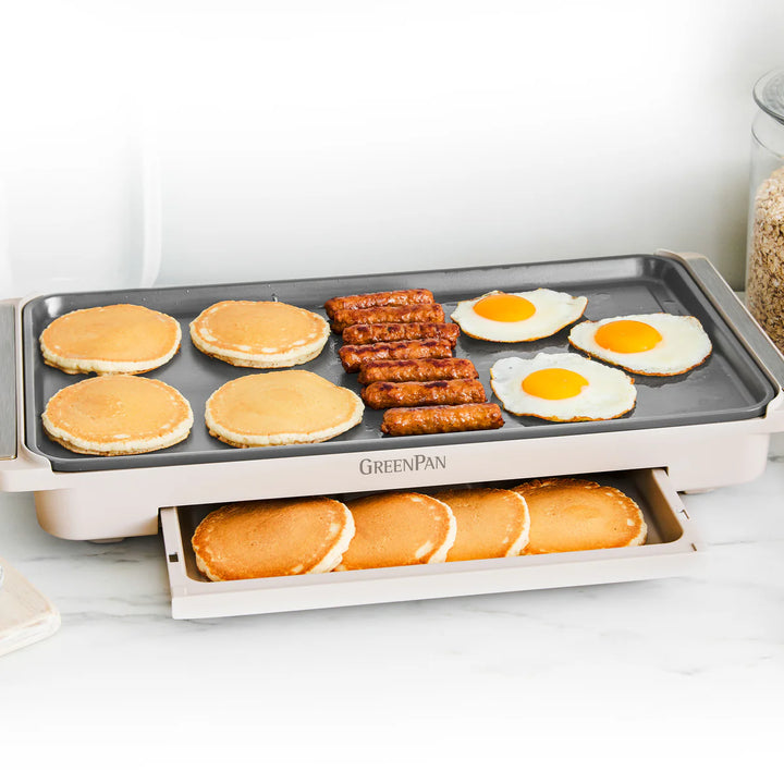Multifunctional griddle with warming drawer with pancakes, eggs, and sausages on a white surface.