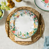 A round plate with floral design sitting on a rattan charger with gold fork and knife on a neutral table cloth.