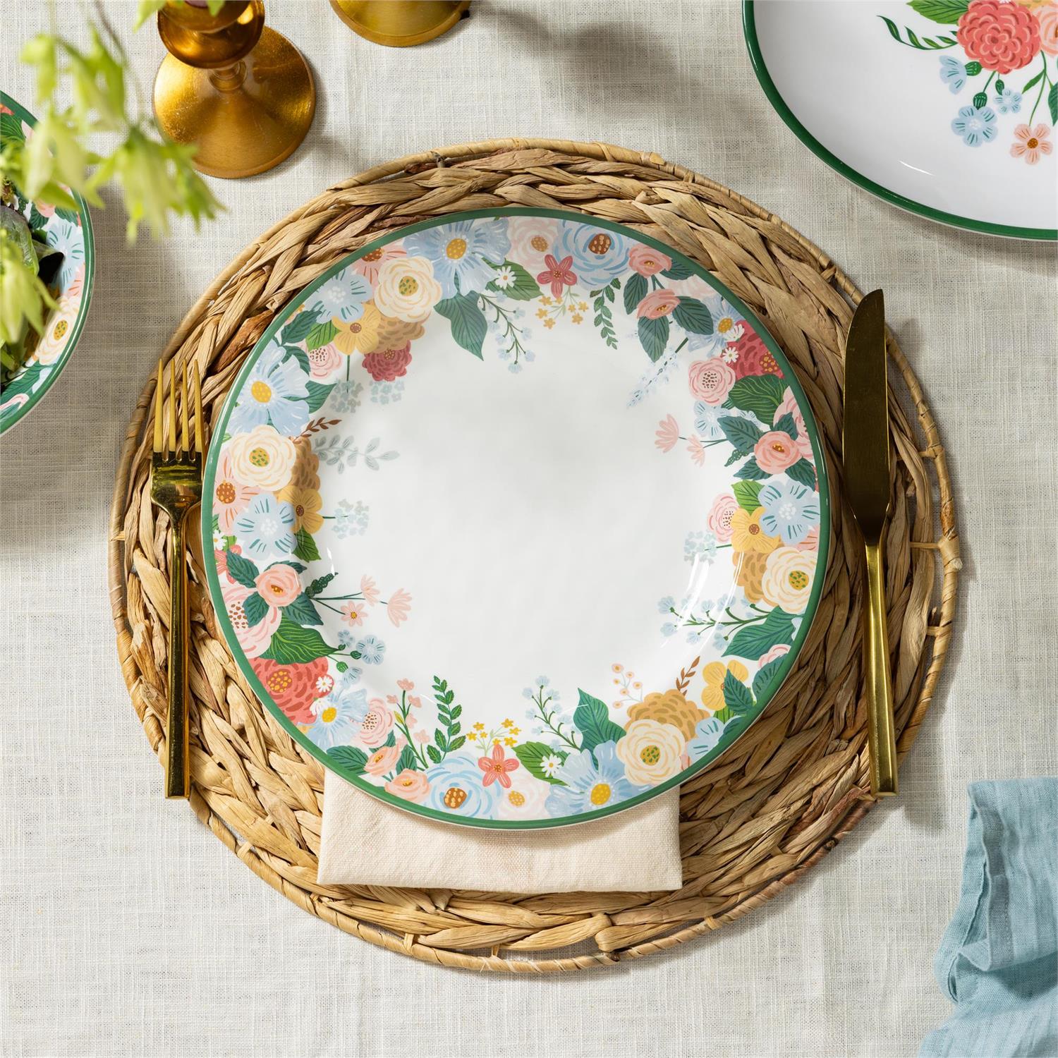 A round plate with floral design sitting on a rattan charger with gold fork and knife on a neutral table cloth.