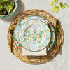 A table setting featuring two floral design plates, gold fork and knife on a  rattan charger; a bowl of salad in the background.