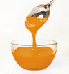 Orange frosting being poured from a spoon into a glass bowl on a white background