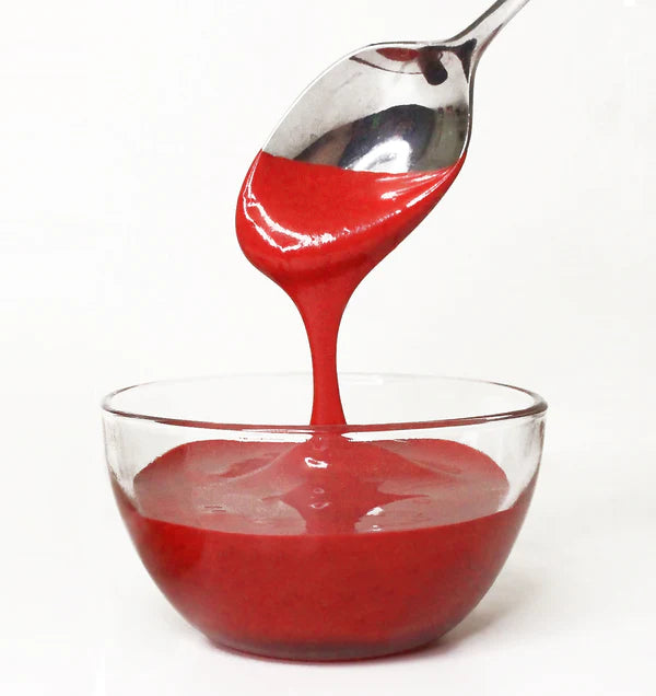 Red frosting being poured from a spoon into a clear glass bowl on a white background