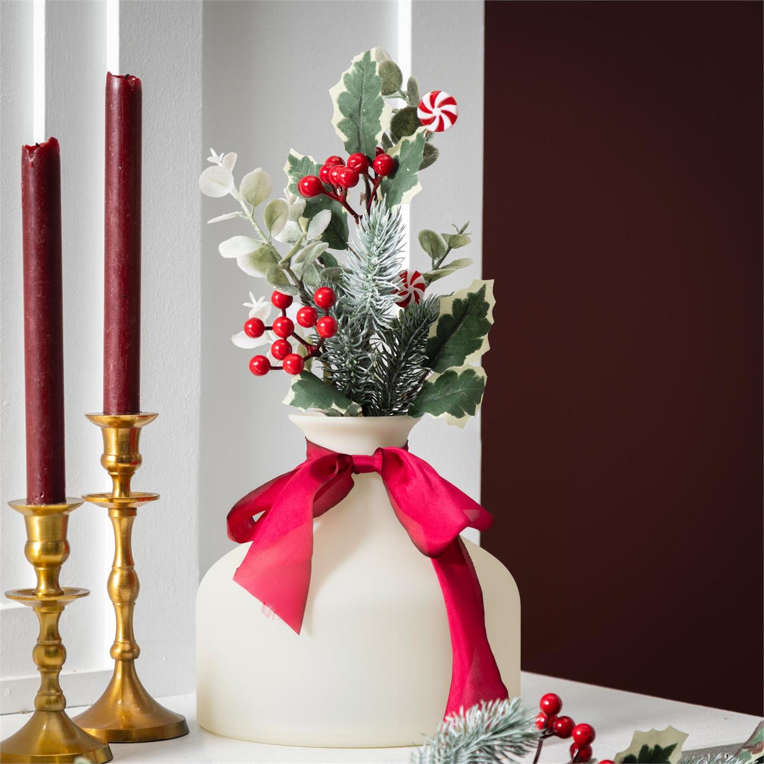 Decorative Christmas greenery stem including berries, holly, and a red ribbon, set against a neutral background.