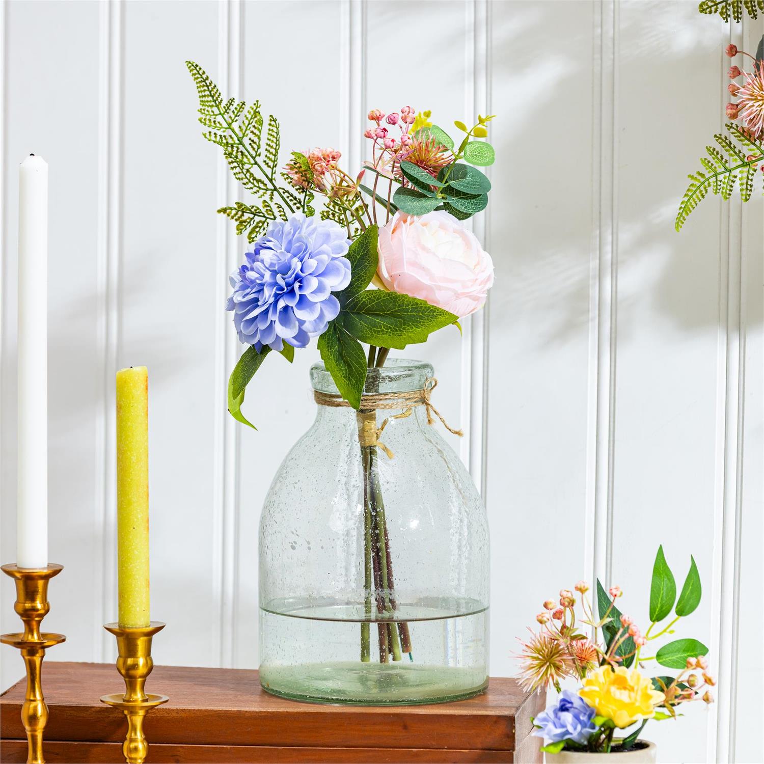 A vase of flowers sitting on a wood bench with candles in the foreground with a white background.