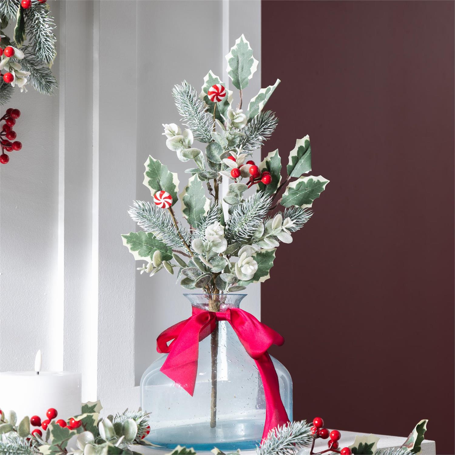 Decorative arrangement with greenery, red berries, and a red ribbon in a clear glass vase against a neutral background.